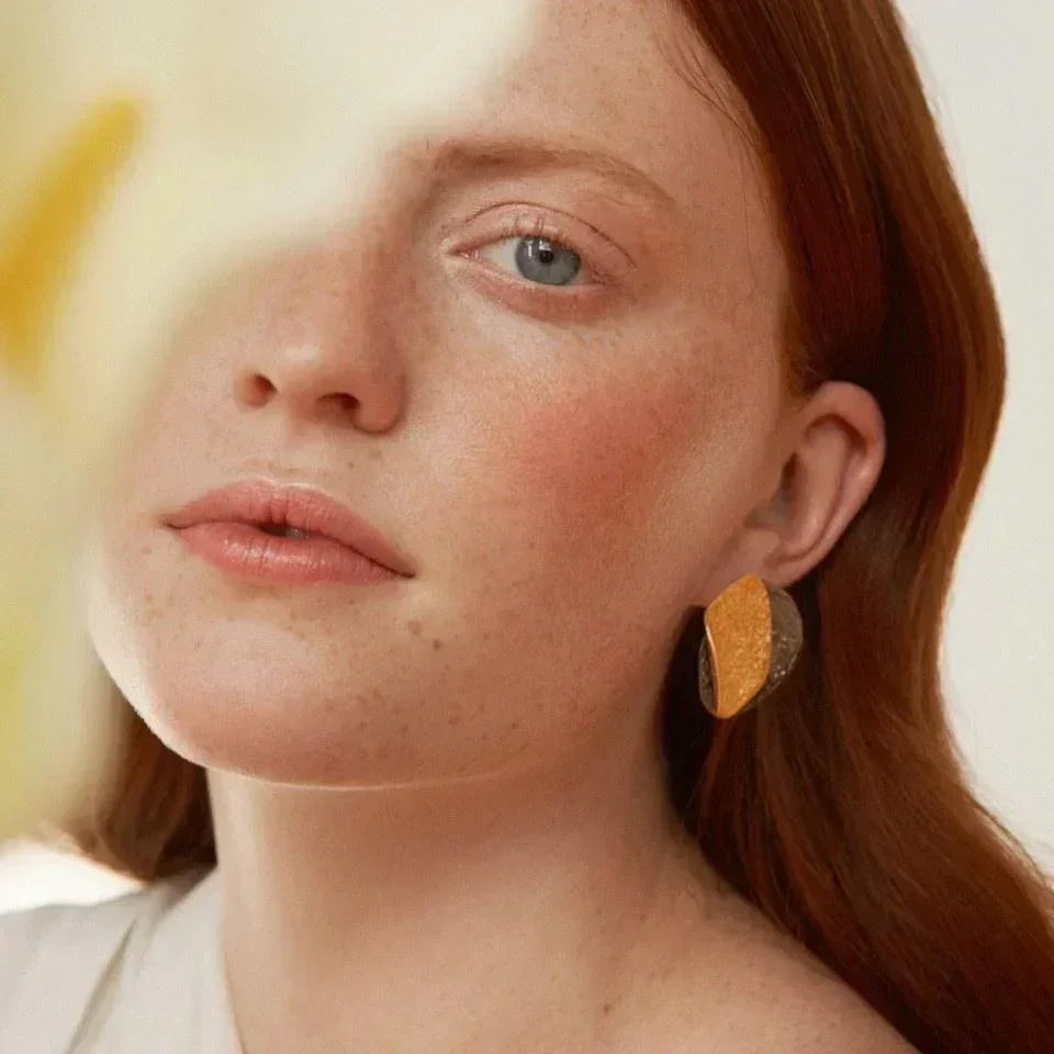 Woman wearing large gold-plated statement earring, soft natural light, close-up jewelry shot