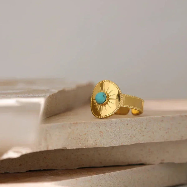 18K gold-plated ring with sunburst design and round turquoise gemstone on stone surface