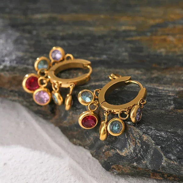 Gold-plated hoop earrings with colorful gemstone charms on a stone background