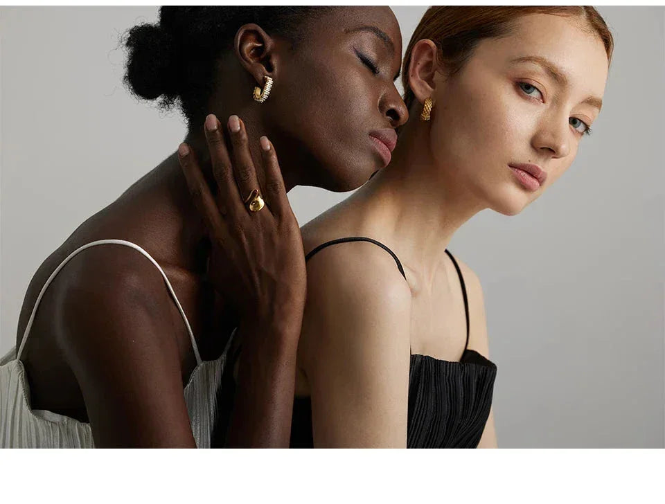 Two women modeling elegant 18K gold-plated earrings and a gold ring in a minimal studio setting.