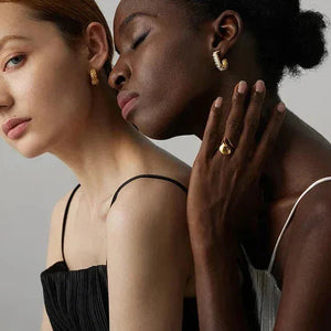 Two women modeling elegant 18K gold-plated earrings and a gold ring in a minimal studio setting.