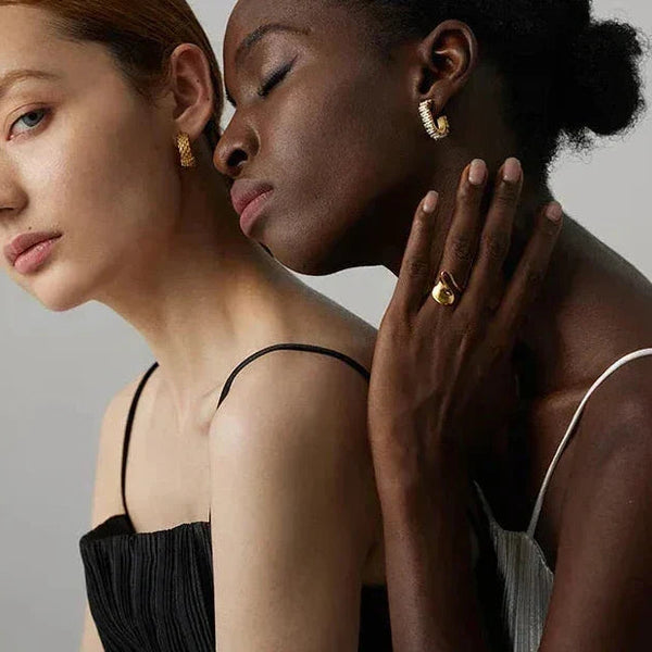 Two women modeling elegant 18K gold-plated earrings and a gold ring in a minimal studio setting.