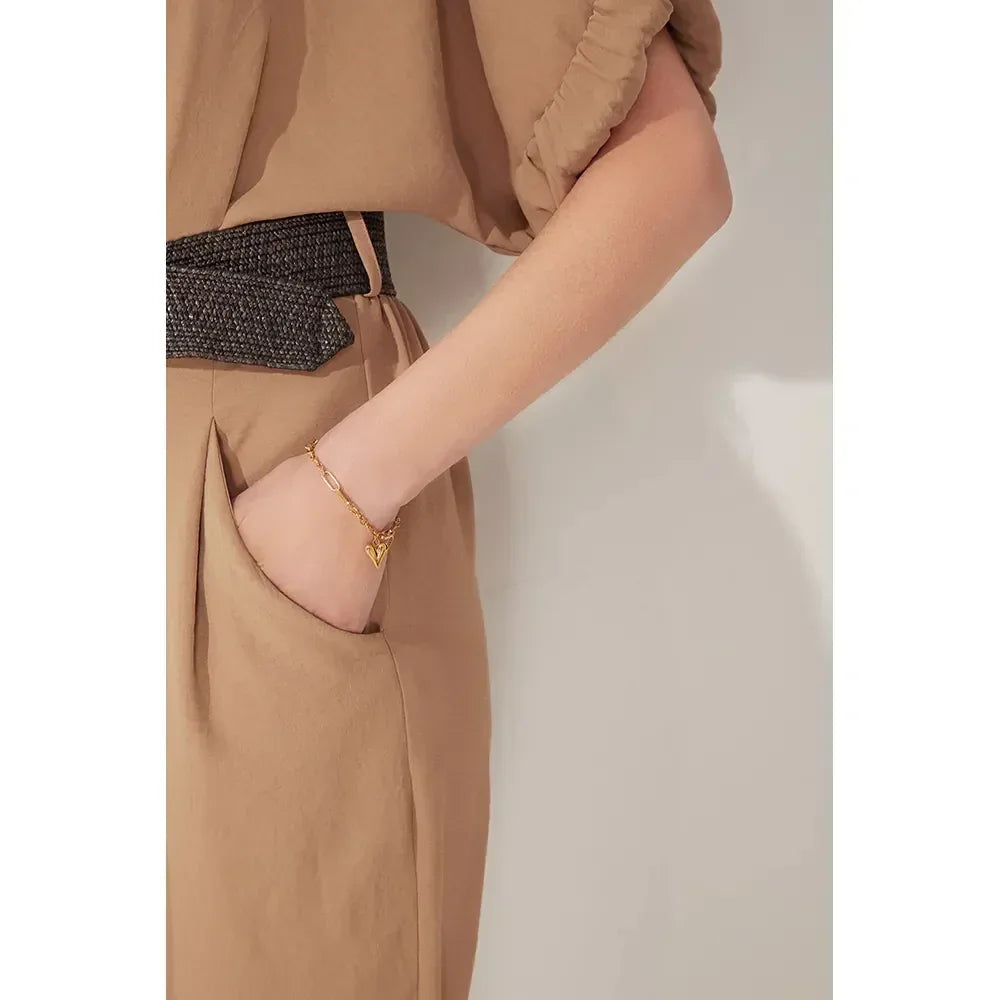 Woman wearing a gold-plated heart charm bracelet with hand in pocket, neutral outfit