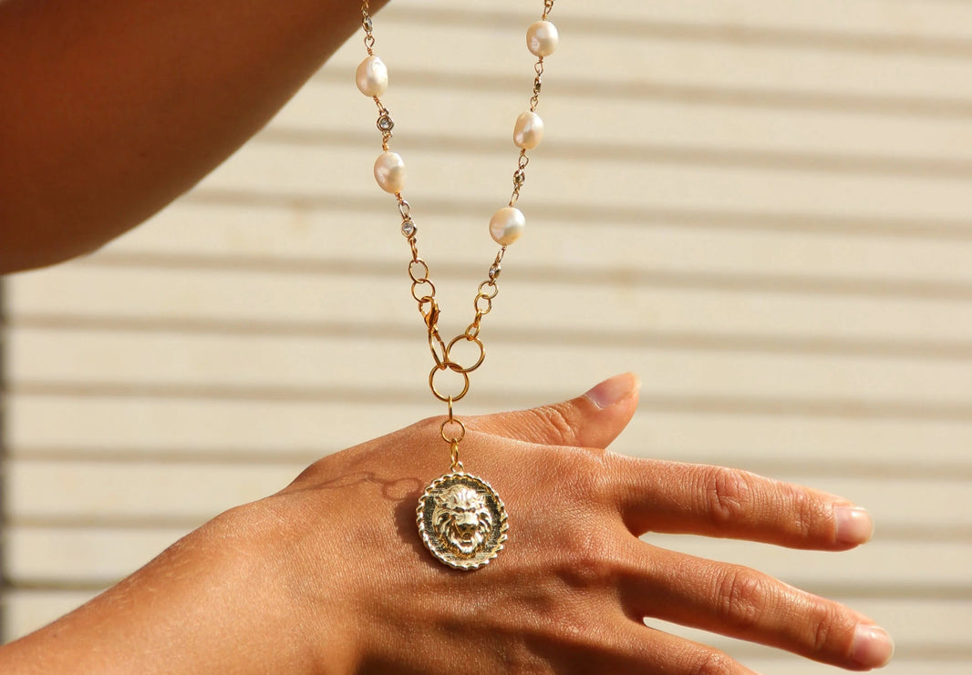 Hand wearing a gold necklace with a pendant against a neutral background