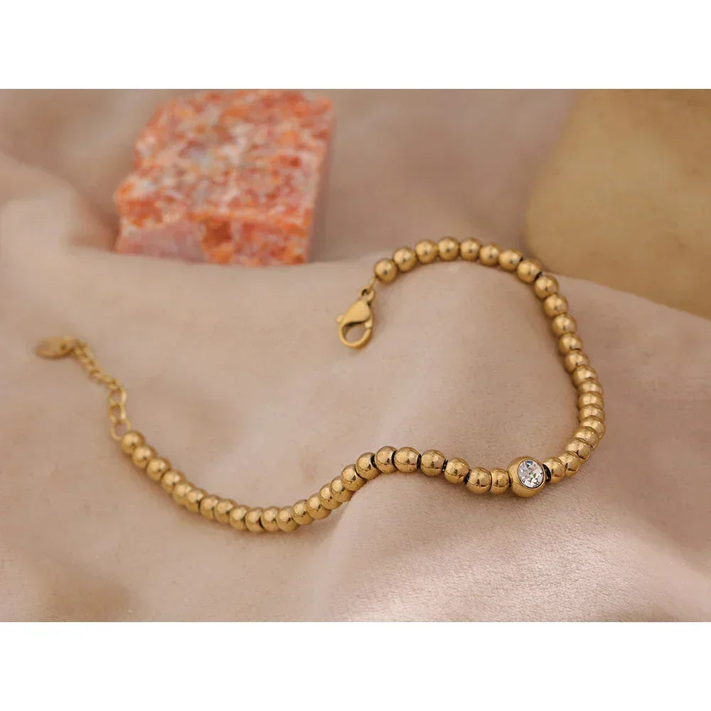 18K gold-plated beaded bracelet with round crystal accent on a soft beige fabric background