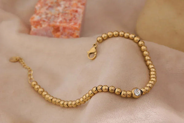 18K gold-plated beaded bracelet with round crystal accent on a soft beige fabric background