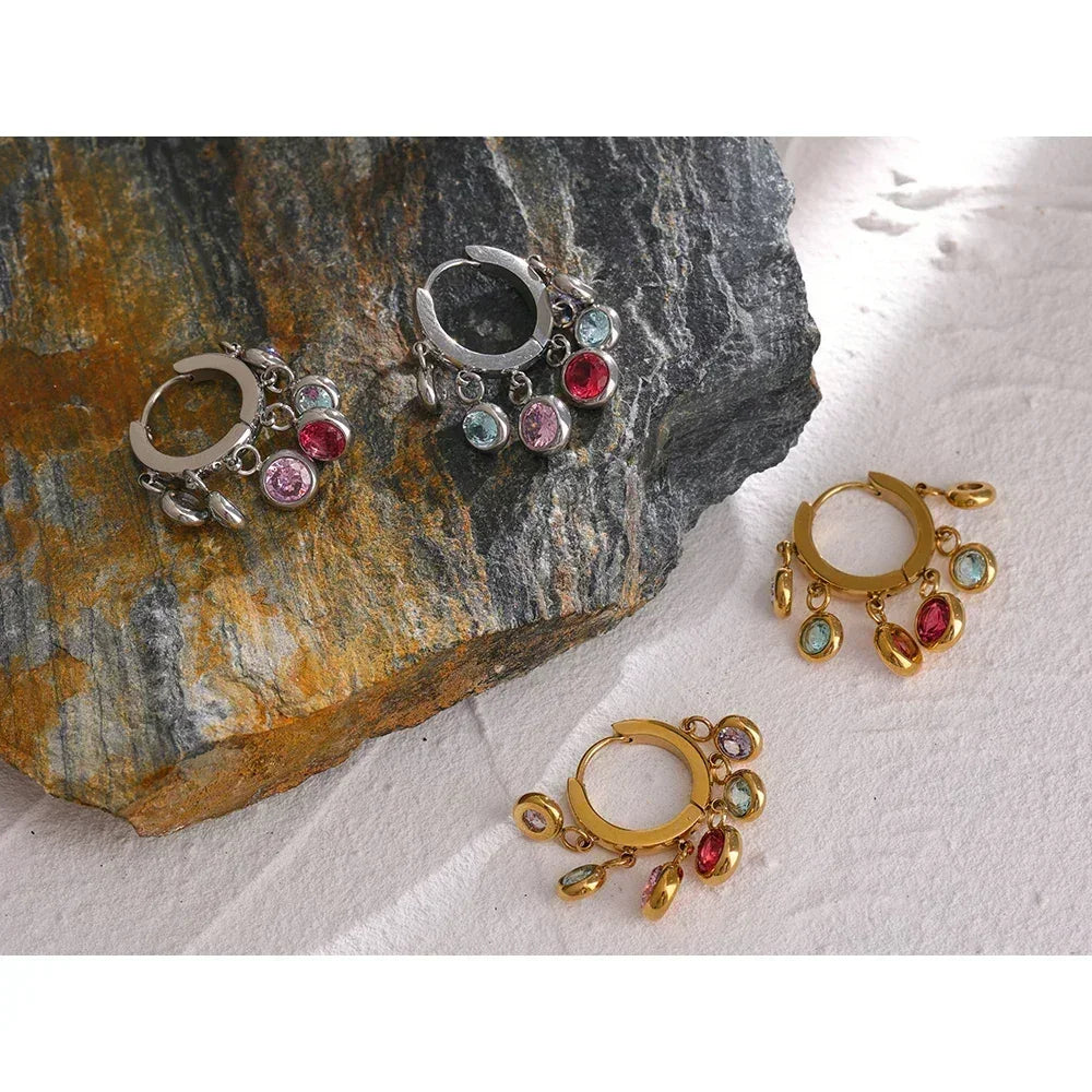 Silver and gold hoop earrings with colorful gemstone charms on textured stone and fabric