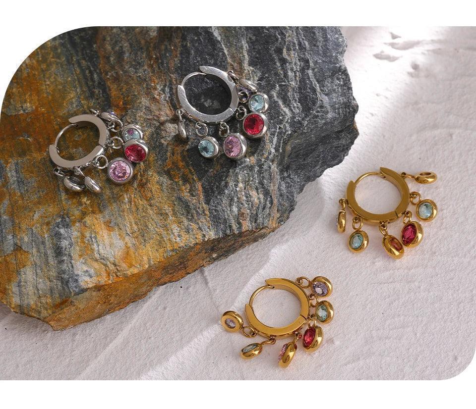 Gold and silver hoop earrings with colorful gemstone charms on textured stone and sand