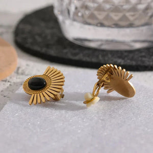 Gold earrings with black and white stones on a textured surface with a glass in the background.