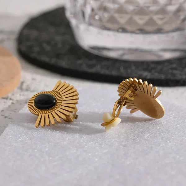 Gold earrings with black and white stones on a textured surface with a glass in the background.
