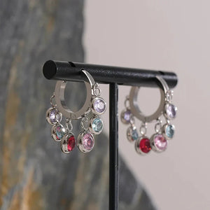 Silver hoop earrings with colorful gemstones on a black stand against a marble background