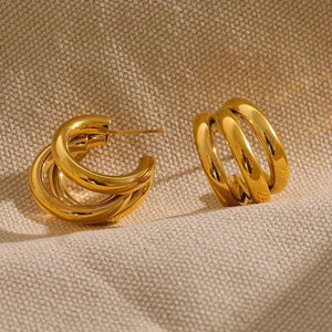 Chunky gold-plated hoop earrings with a triple band design on beige textured fabric.