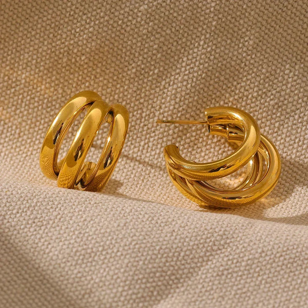 Chunky gold-plated hoop earrings with a triple band design on beige textured fabric.