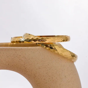 Pair of hammered 18K gold-plated bangles displayed on a curved ceramic stand