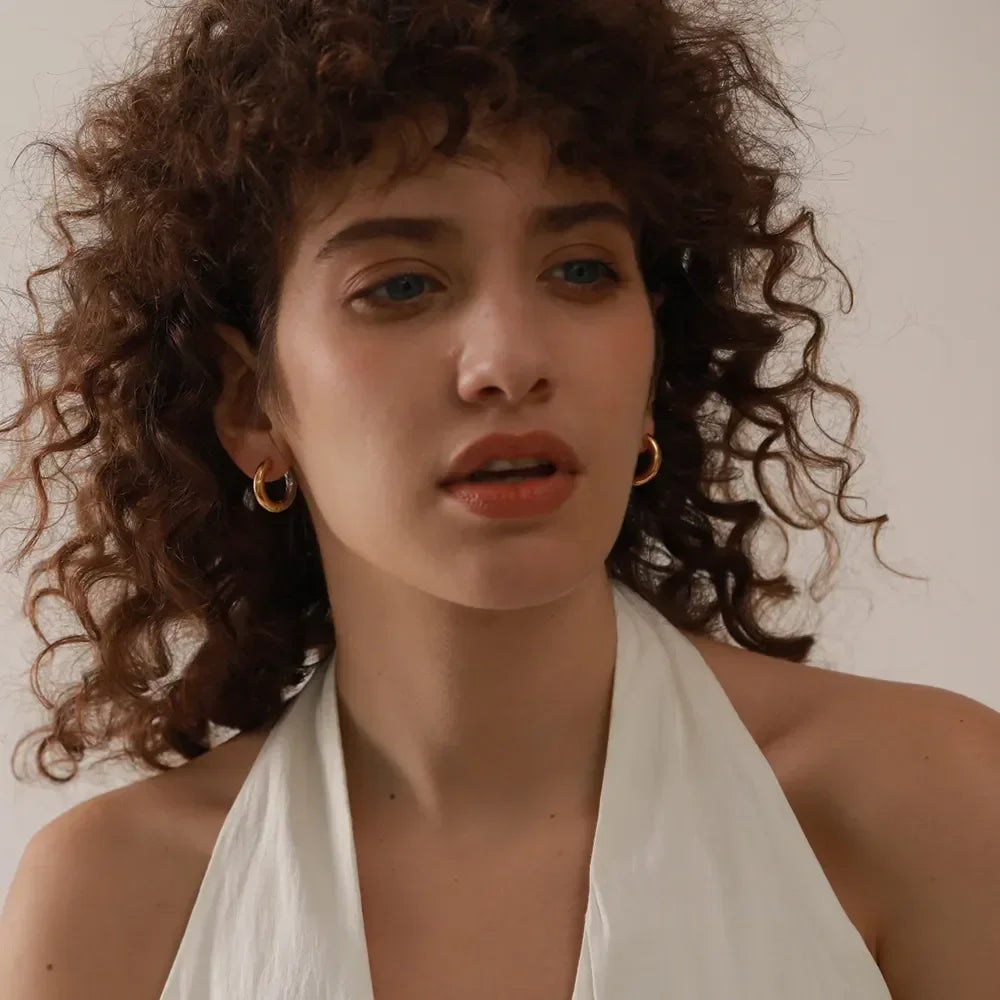 Woman wearing 18K gold-plated hoop earrings and a white halter top, curly hair, neutral background