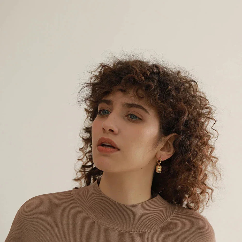 Woman wearing gold-plated dangle earrings and taupe top, minimalist background