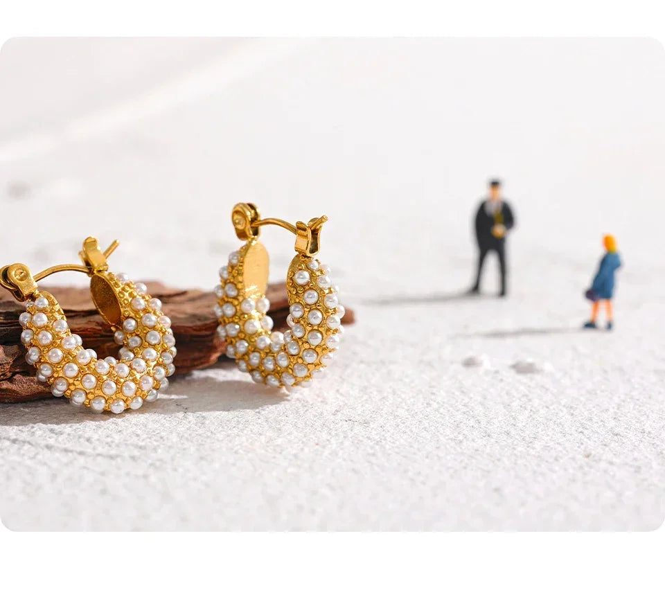 Gold-plated hoop earrings with white pearls on textured surface, miniature figures in background