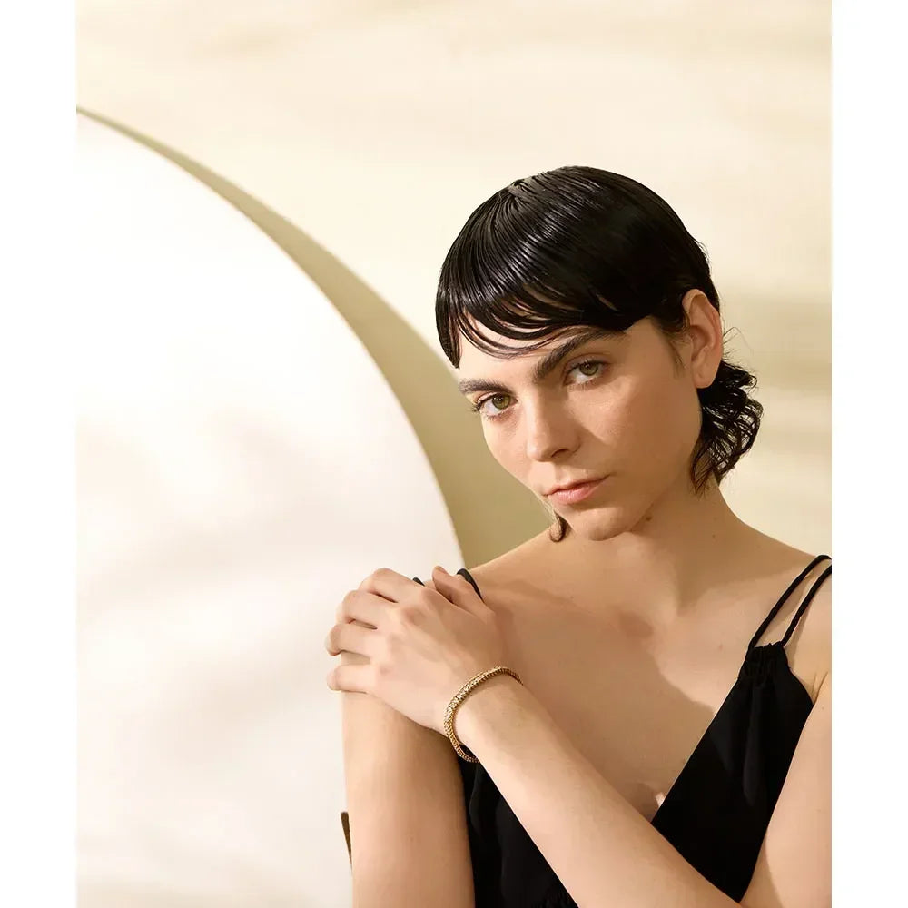 Woman wearing gold-plated bracelet and black dress, posing against neutral background