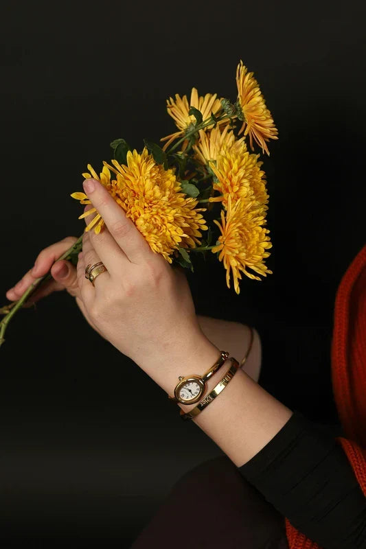 Hand holding a bouquet of yellow flowers with a gold watch and ring on a dark background